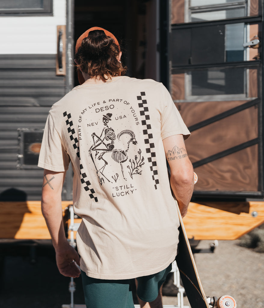 A man wearing the Still Lucky Tee in bone variant by Deso Supply Co. from the back view.