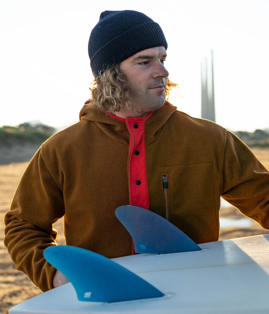 A man with a surf board wearing Tenaya Snap Hoodie in pecan w/ rufous variant by Deso Supply Co. 1