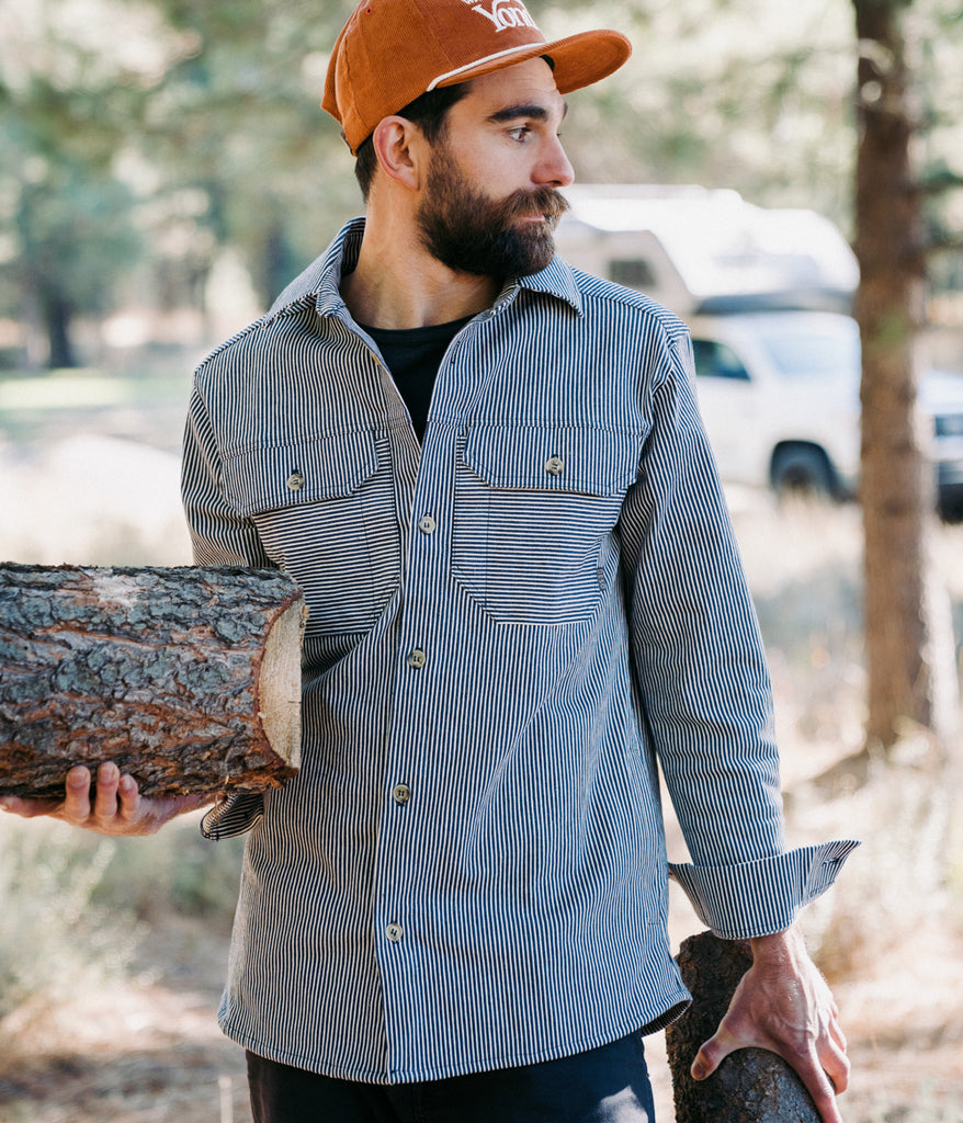 A man, dressed in an Indigo Dyed Workshop Shirt by Deso Supply Co., is holding a log outdoors.