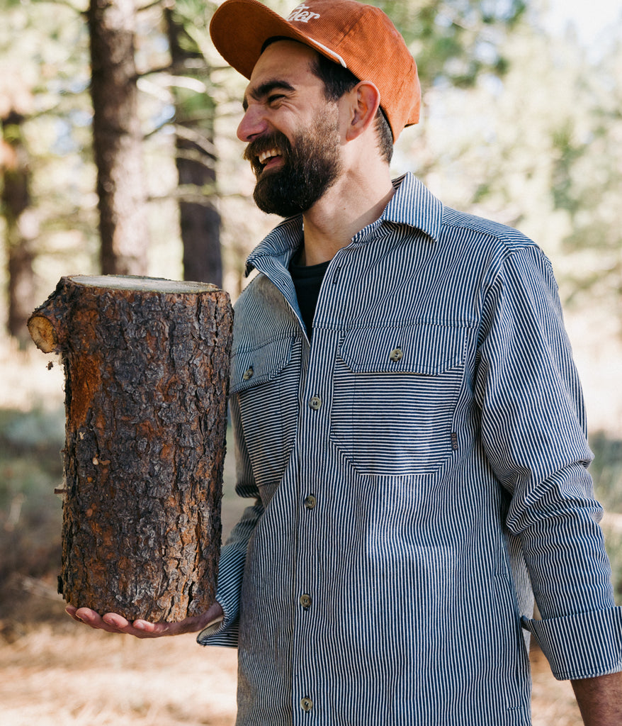 A man dressed in an Indigo Dyed Workshop Shirt by Deso Supply Co., is holding a log outdoors. 1