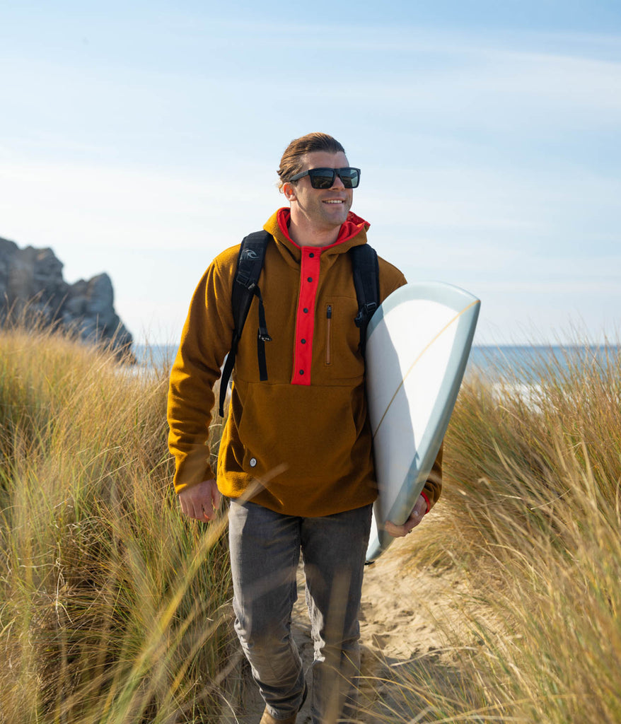 A man with a surf board wearing Tenaya Snap Hoodie in pecan w/ rufous variant by Deso Supply Co.