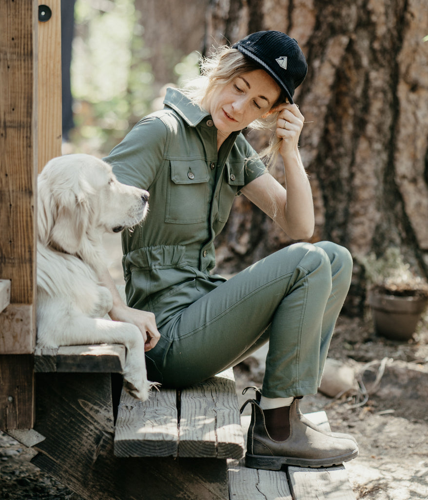 A woman beside a dog wearing Homewood Coveralls in dark sage color by Deso Supply Co.